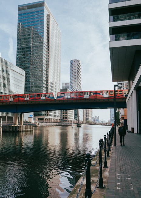 Exterior view of Canary Wharf Station in daylight, featuring a modern glass and metal entrance canopy with the London Underground roundel sign visible. Surrounding the station are tall office building