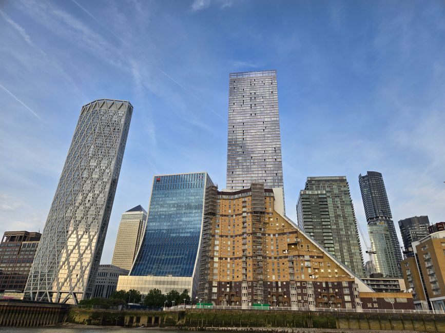 A wide view of the Canary Wharf skyline featuring several modern high-rise office buildings with reflective glass facades, including a distinctive twisted steel structure on the left and a tall, recta