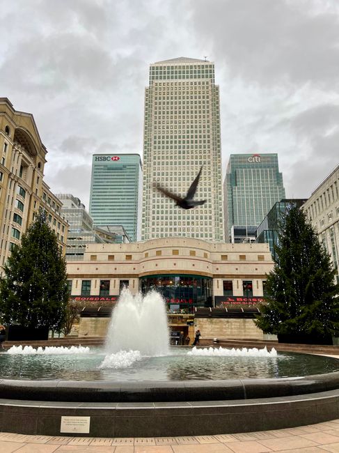 A low-angle view of modern commercial office buildings in Canary Wharf, featuring glass facades reflecting the sky and neighboring structures. The central skyscraper has a white grid-like exterior, wh