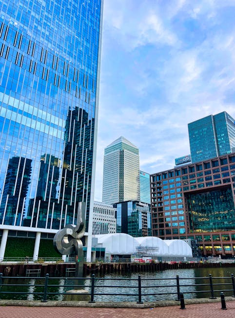 Aerial view of Canary Wharf at sunset showcasing a cluster of modern skyscrapers with reflective glass facades and varying architectural styles. The sunlight creates a warm glow on the building exteri